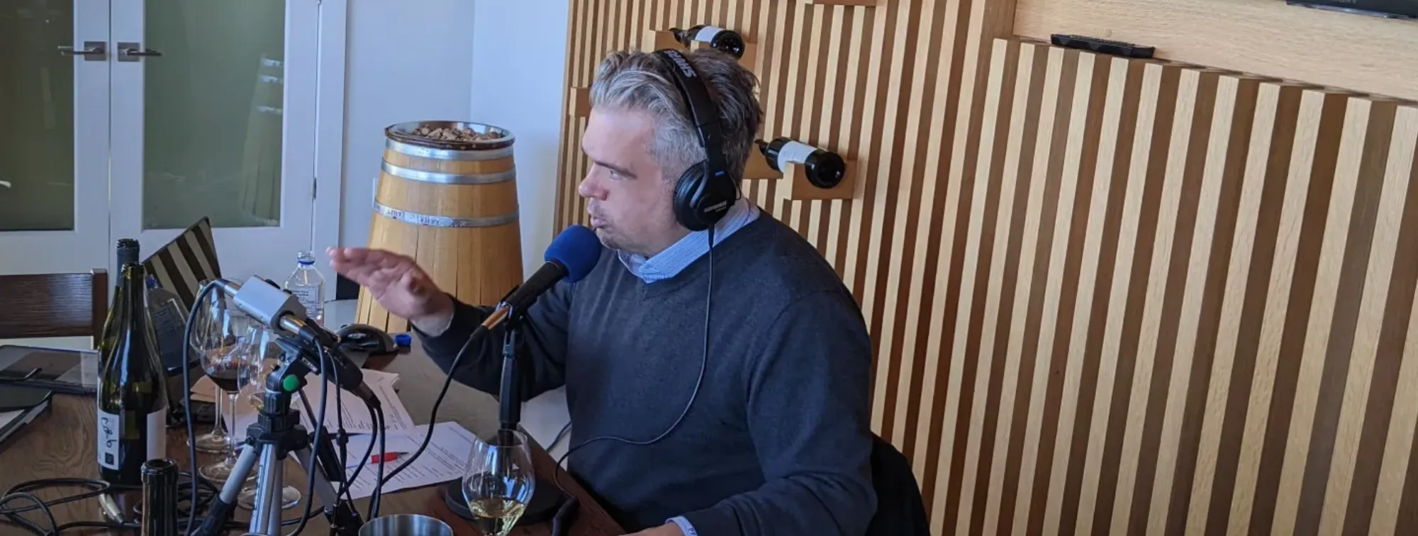 Podcast host speaking into a microphone with wine bottles and glasses on the table, featured on ‘Read Between the Wines’ podcast.